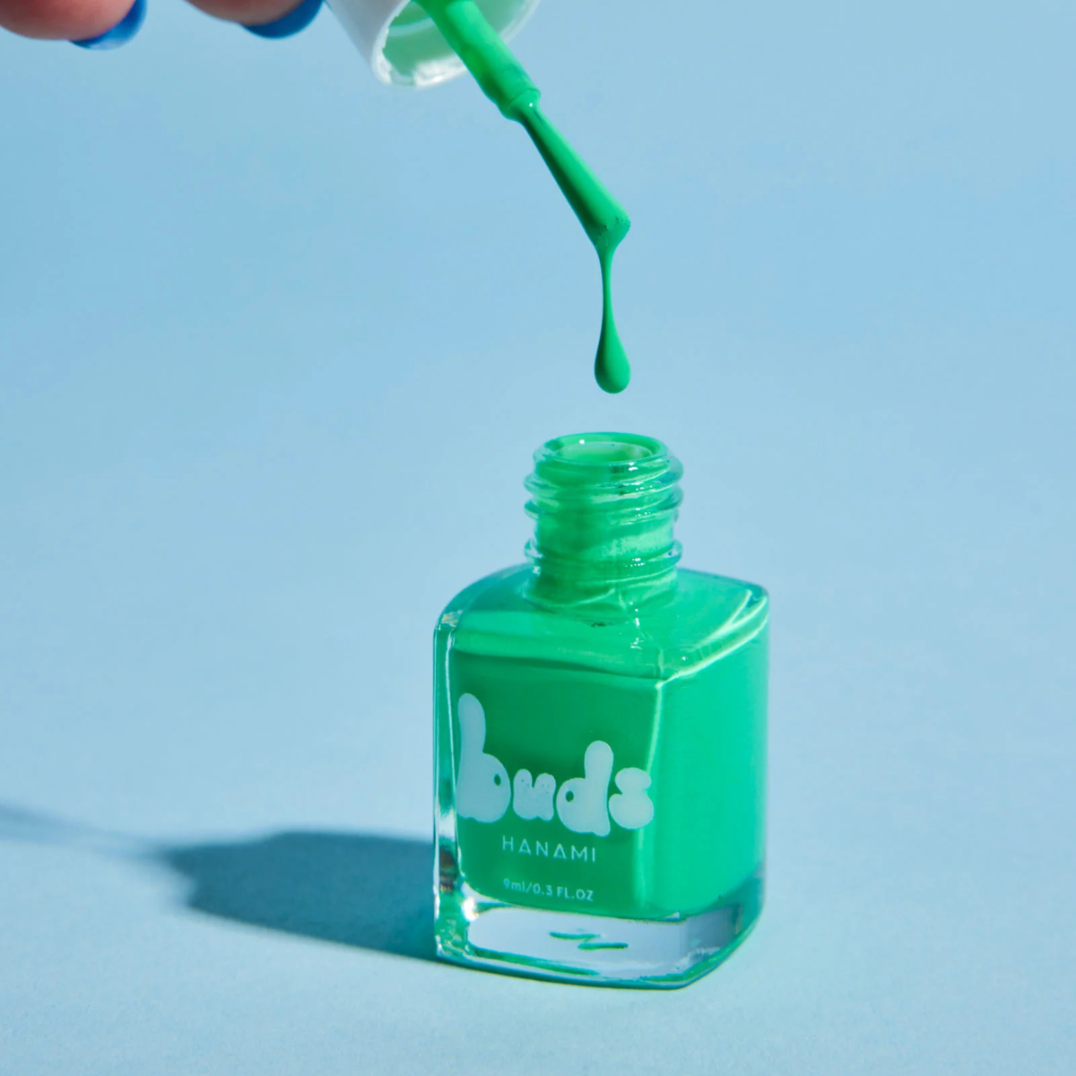 Green Buds kids nail polish bottle with the brush dripping polish on a blue background